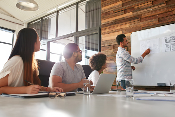 Trainer standing at whiteboard with delegates looking on and taking notes.