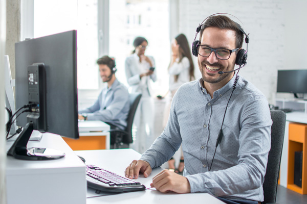 Smiling male support desk worker wearing headphones with colleagues in background.